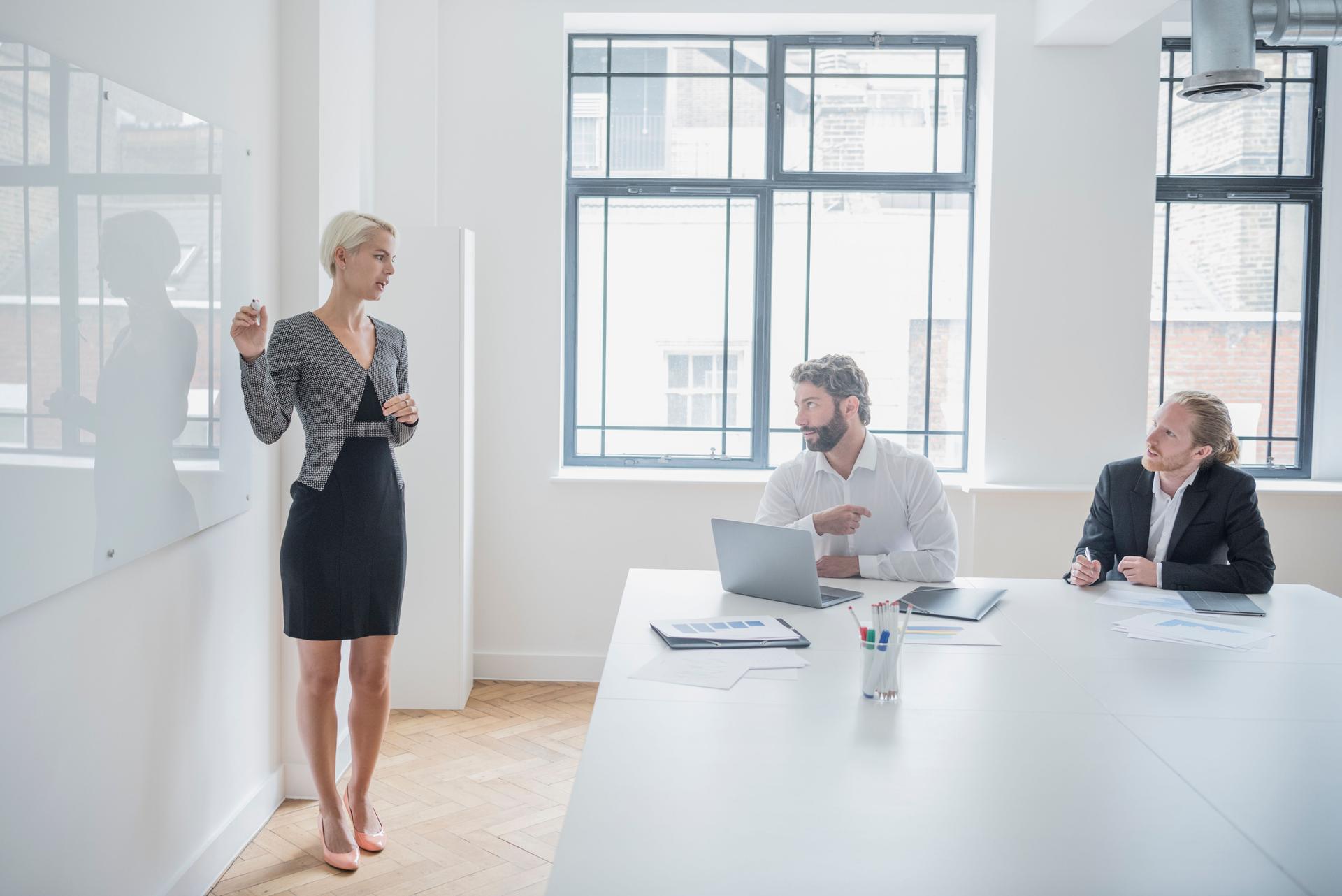 Businesswoman standing at whiteboard giving presentation, two male colleagues listening